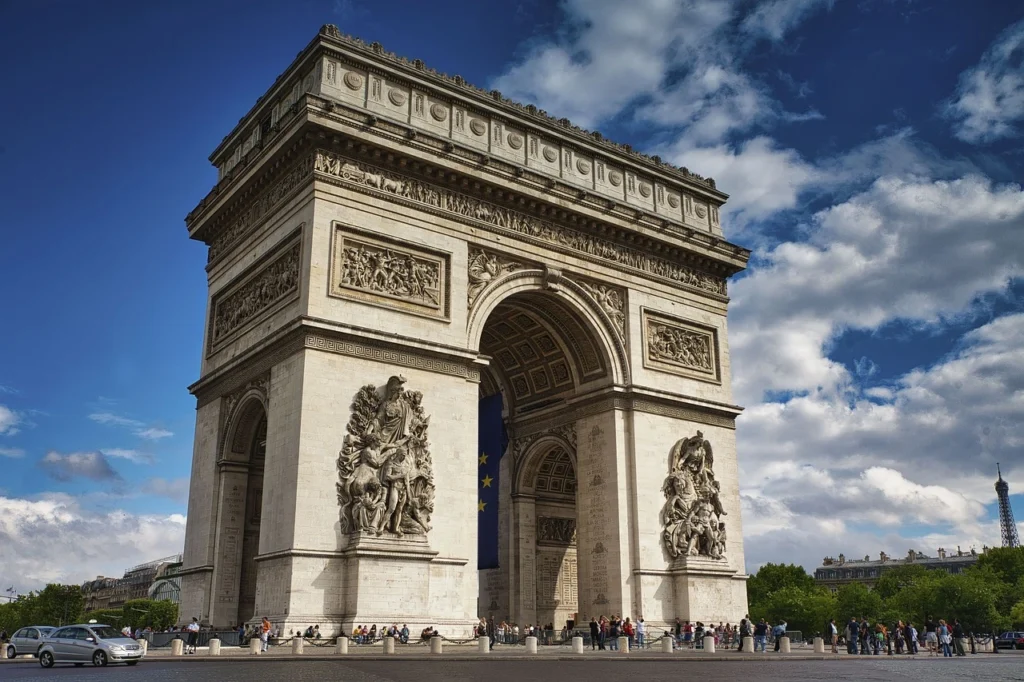 Güneşli bir günde, Paris'in kalbinde yükselen, detaylı kabartmaları ve görkemli kemerleriyle dikkat çeken Zafer Takı'nın çevresinde gezinen ziyaretçiler ve uzakta belirginleşen Eyfel Kulesi ile şehir, Her Köşesi Bir Film Sahnesi: Paris Gezi Rehberi için büyüleyici bir an sunuyor.