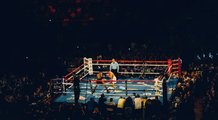 Karanlık bir arenada parlak ışıklarla aydınlatılmış boks ringinde, bir hakem gözetiminde kıyasıya mücadele eden iki boksör ve onları dikkatle izleyen kalabalık seyirciler, 2024 Olimpiyatları'na katılacak Türk sporcuların heyecanını ve azmini yansıtan bir an.