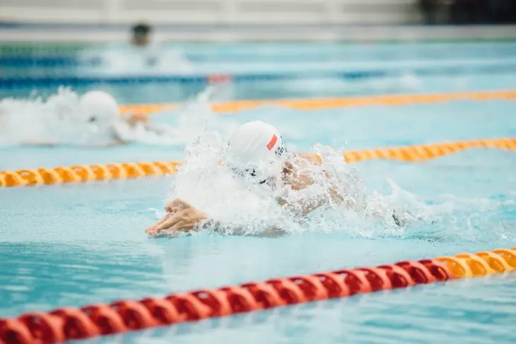 Beyaz boneli bir yüzücü, 2024 Olimpiyatları'nın ruhunu yansıtan bir anla, turkuaz renkteki bir yarış havuzunda güçlü kulaçlar atarak yoğun su sıçramaları yaratıyor; ön planda kırmızı ve turuncu kulvar çizgileri belirginleşirken, arka planda bulanık bir diğer sporcu da mücadele ediyor.