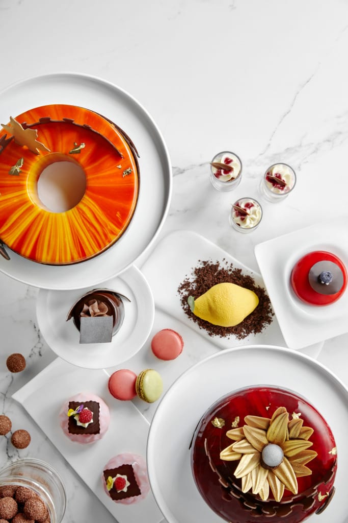 Lüks bir Raffles brunch'ında sunulan, turuncu parlak soslu halka pasta, sarı limon şeklindeki tatlı ve çikolata kırıntıları, kırmızı jöleli ve altın çiçek detaylı pasta, renkli makaronlar ile kremalı ve meyveli küçük bardak tatlıları gibi göz alıcı ve zarif çok sayıda lezzet beyaz mermer bir yüzeyde şıkça sergileniyor.