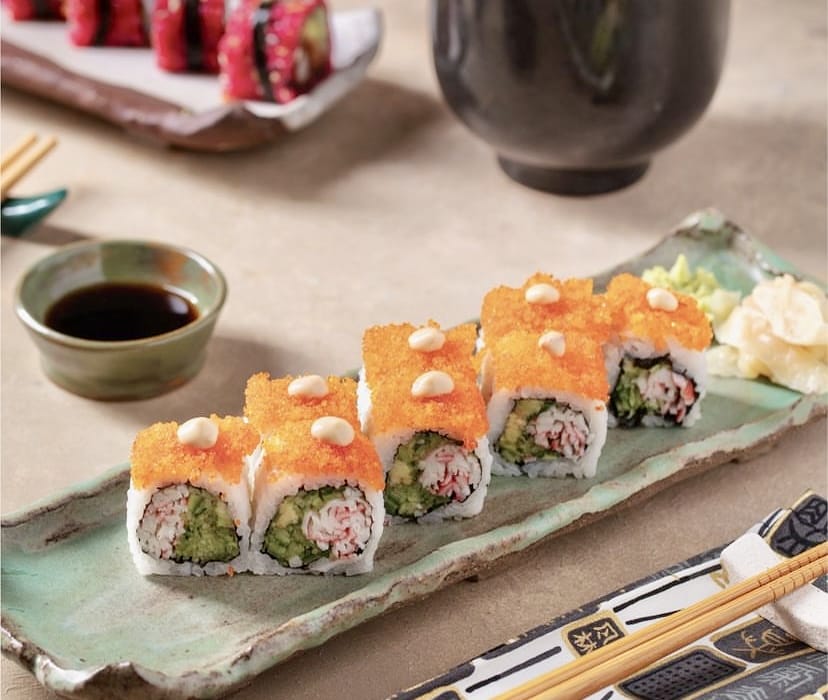 Üzeri parlak turuncu tobiko ve beyaz sos damlacıklarıyla kaplı, içinde yengeç ve avokado bulunan dilimlenmiş uramaki sushi rulolarının yeşil seramik bir tabakta, yanında taze wasabi ve zencefil turşusu ile sunulduğu bu görsel, İstanbul’un en popüler sushi restoranlarının özenli sunumlarını yansıtıyor.
