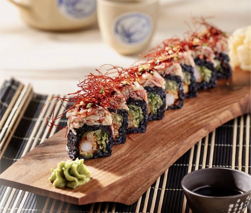 Ahşap bir servis tahtası üzerinde, siyah pirinçle hazırlanmış, üzeri kırmızı biber ipleri ve deniz mahsulleriyle zenginleştirilmiş altı adet özel uramaki sushi, yanında yeşil wasabi ve soya sosu ile İstanbul'un rafine sushi restoranlarında sunulan modern bir lezzeti vurguluyor.