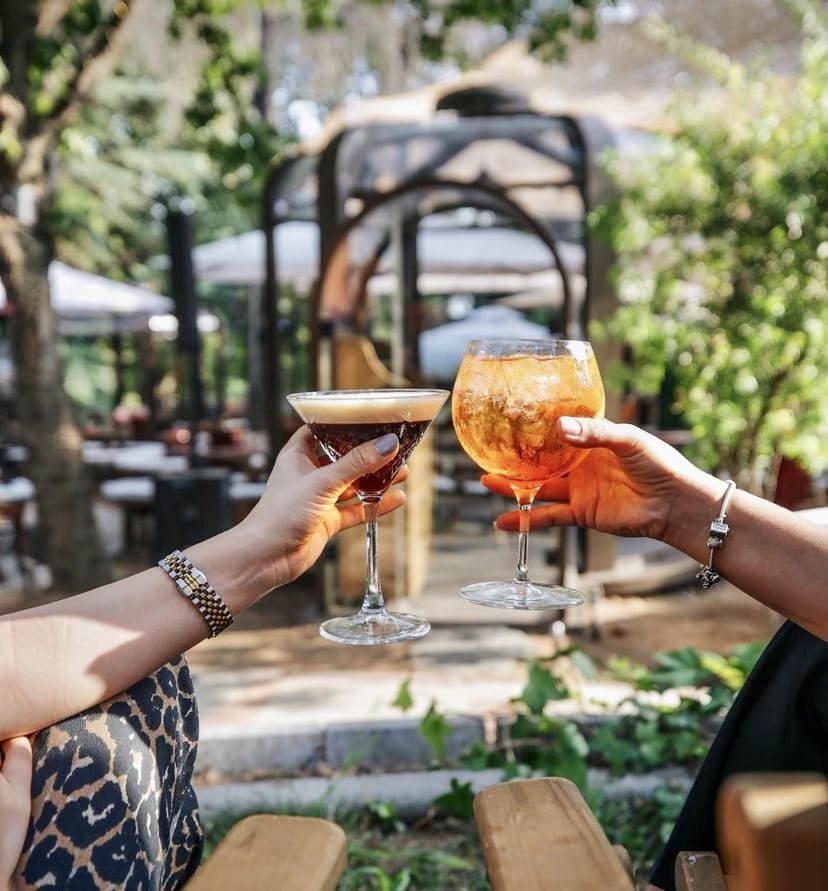 İki kişi, yemyeşil ve huzurlu bir bahçeli mekânın ferah atmosferinde kadeh kaldırarak, birinde koyu renk köpüklü martini bardağı, diğerinde ise buzlu Aperol Spritz bardağı tutarken, İstanbul'un en güzel bahçeli mekânlarından birinde keyifli bir anın tadını çıkarıyor.