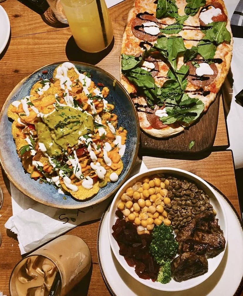 Ahşap bir masada üstten çekilmiş bir karede, üzerinde roka, salam ve peynir olan dikdörtgen bir pizza, guacamole ve beyaz sosla zenginleştirilmiş nachos tabağı, nohut, mercimek, brokoli, pancar ve ızgara et parçacıkları içeren sağlıklı bir kase, buzlu kahve ve limonata, İstanbul'un bahçeli mekanlarında sunulan zengin lezzet çeşitliliğini gözler önüne seriyor.