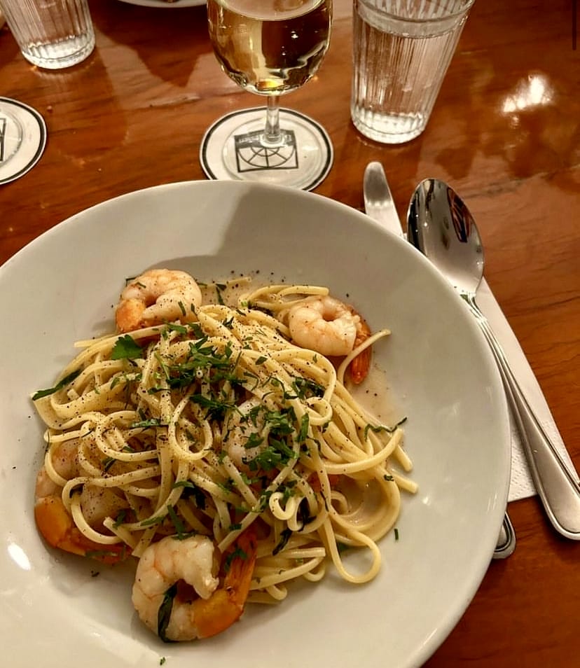 Ahşap bir masada, ince spagetti veya linguine ile hazırlanmış, bol karidesli, maydanoz ve karabiber serpilmiş lezzetli bir makarna tabağı, yanında bir kadeh beyaz şarap ve su bardağıyla, İstanbul'daki kaliteli mekanlarda sunulan seçkin bir yemeği temsil ediyor.