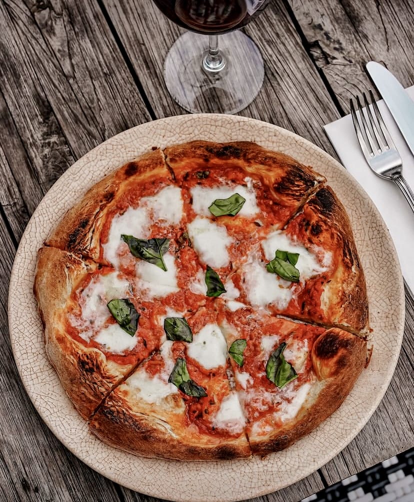 Ahşap bir masada, dilimlenmiş, üzerinde erimiş mozzarella peyniri ve taze fesleğen yaprakları bulunan nefis bir Napoli pizzası, yanında bir kadeh kırmızı şarap ve çatal bıçak takımıyla İstanbul'un bahçeli mekanlarında keyifli bir açık hava yemeği deneyimini çağrıştırıyor.