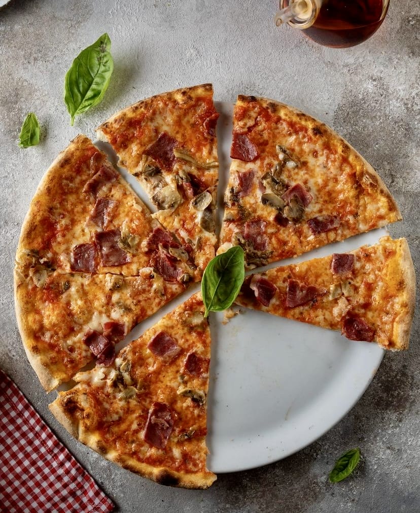 Beyaz bir tabakta servis edilmiş, üzerindeki sucuk, mantar ve fesleğen yapraklarıyla iştah açıcı görünen, altı dilime ayrılmış lezzetli bir pizza, İstanbul'daki keyifli bahçeli mekanlarda sunulan zengin menülerin cazibesini vurguluyor.