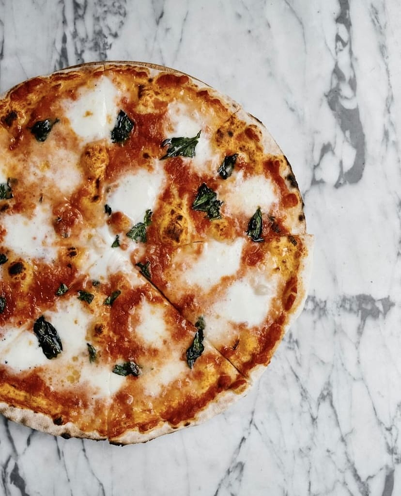 Mermer bir zeminde duran, üzerinde bolca erimiş mozzarella peyniri, canlı kırmızı domates sosu ve taze yeşil fesleğen yaprakları bulunan, birkaç dilimi ayrılmış iştah açıcı Napoli usulü pizza, İstanbul’un en beğenilen bahçeli mekânlarında sunulan gurme lezzetleri vurguluyor.