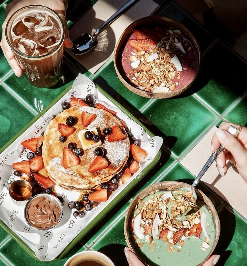 Nove Garden'da yeşil karolu bir masada servis edilen, çilek, yaban mersini ve tereyağı ile süslü pankekler, granolalı iki farklı smoothie kasesi ve buzlu kahve, İstanbul'un en güzel bahçeli mekânları arasında keyifli bir kahvaltı deneyimini yansıtıyor.