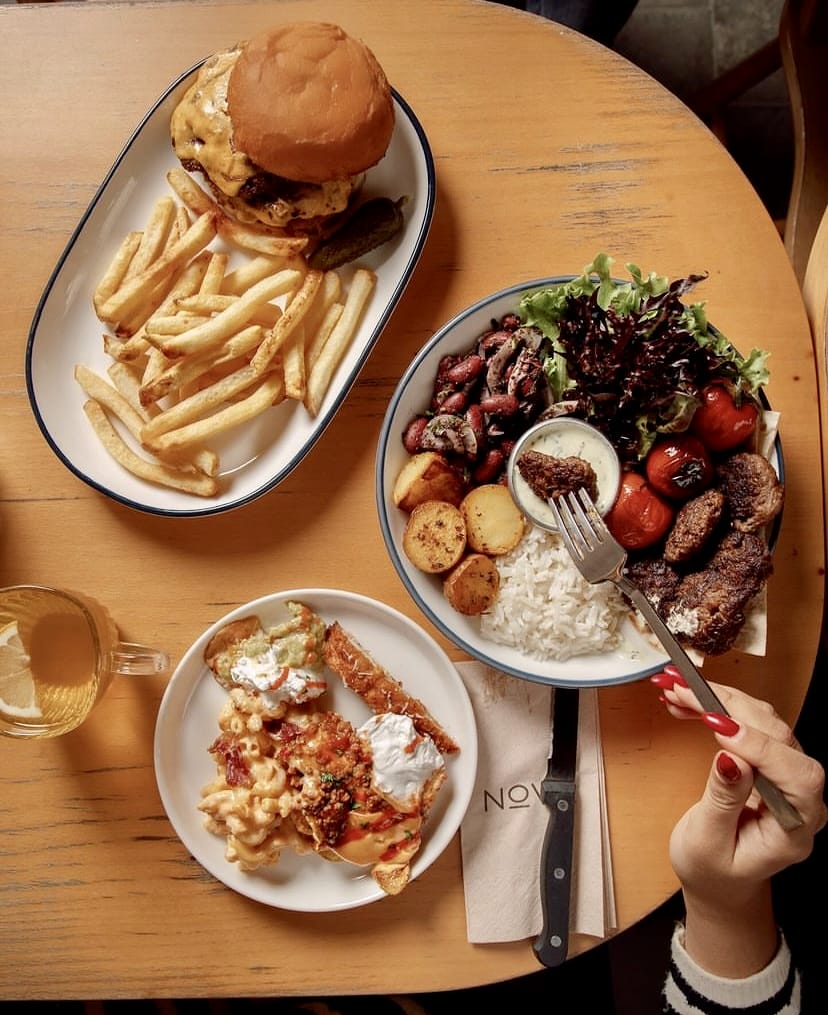 İstanbul'un keyifli bahçeli mekanlarından Nove Garden'da, peynirli burger ve patates kızartması, bol malzemeli bir kase yemek ve kremalı makarnadan oluşan zengin menüden lezzetli seçenekler sunan dolu bir masa.