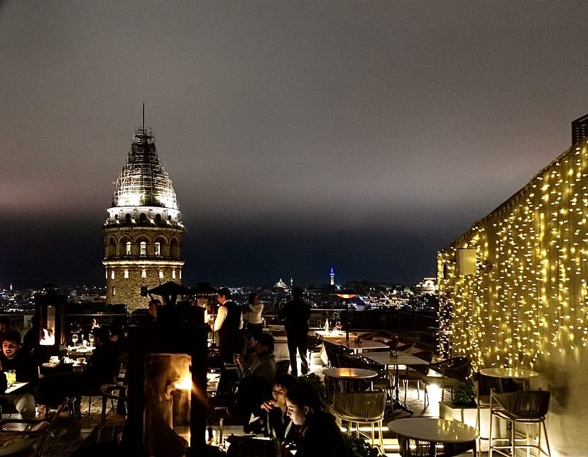 İstanbul'un en iyi teraslı mekanlarından birinde, Galata Kulesi'nin parıldayan görüntüsü eşliğinde, şık bir duvarı süsleyen sarı dekoratif ışıkların altında toplanan misafirler, şehrin büyüleyici gece manzarasının ve canlı atmosferinin tadını çıkarıyor.