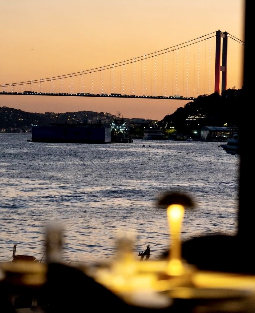 Akşamüstü kızıl gökyüzü altında Boğaziçi Köprüsü'nün üzerindeki araç ışıkları parlarken, Boğaz'ın sakin suları ve ön plandaki bir masada yanan sarı ışıklı lamba, İstanbul'un en iyi teraslı mekanlarından birinde batan güneşe karşı huzurlu bir anı yakalıyor.