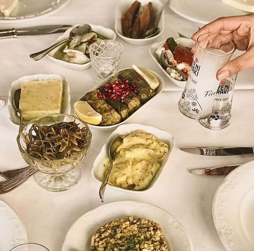 Nar taneleri ve limon dilimleriyle süslenmiş zeytinyağlı sarmaların, çeşitli geleneksel mezelerin ve bir elin tuttuğu rakı kadehinin bulunduğu bu zengin masa, İstanbul'un en iyi teraslı mekanlarında sunulan keyifli bir akşam yemeği deneyimini yansıtıyor.