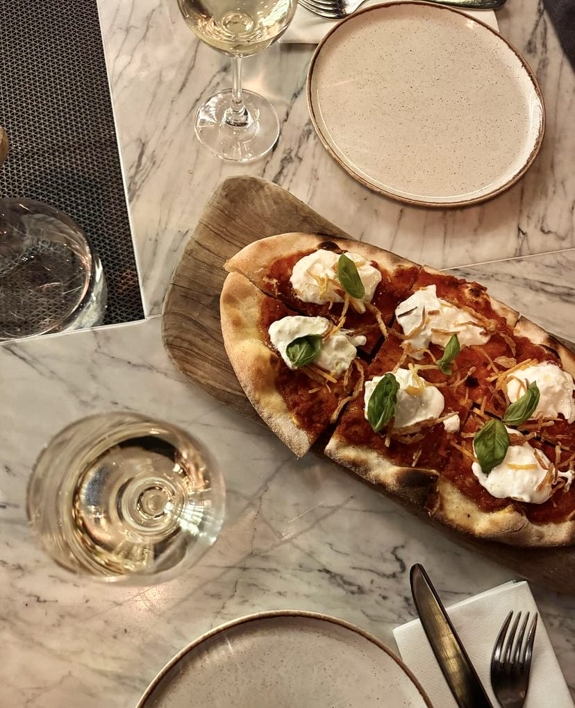 İstanbul'un en iyi teraslı mekanlarından birinde, yaz akşamı keyfiyle beyaz şarap eşliğinde servis edilen, domates soslu, burrata peynirli ve taze fesleğenli uzun bir gurme pizza, mermer bir masada zarif sunumuyla öne çıkıyor.