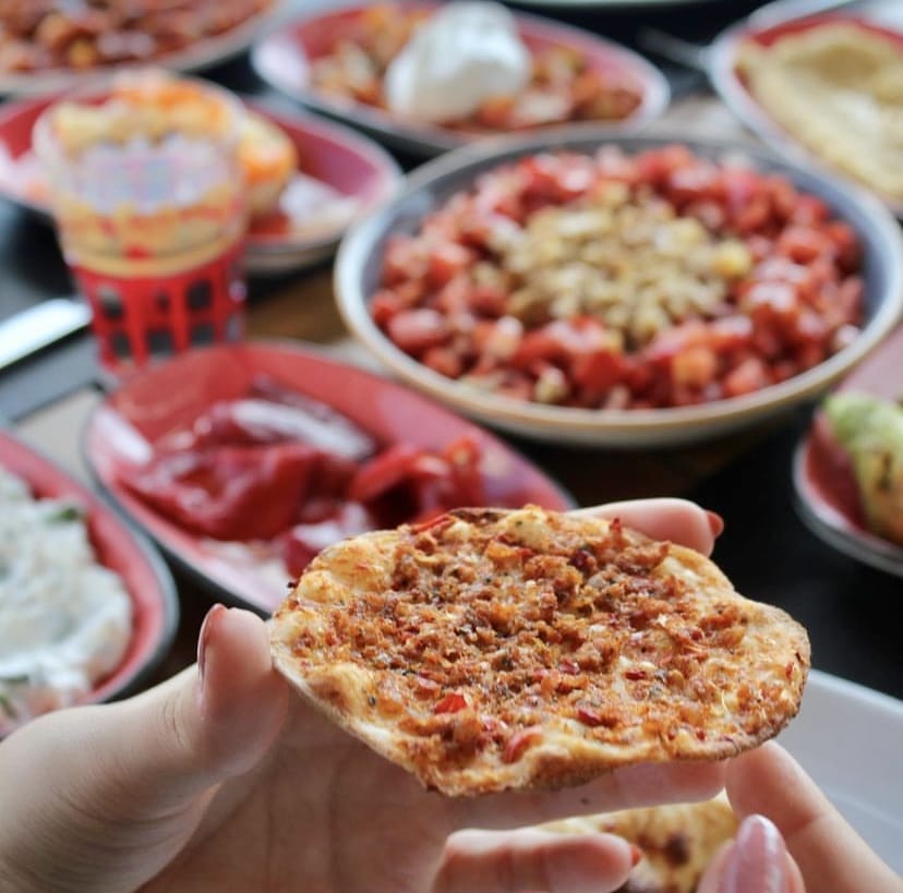 Bir elin tuttuğu lahmacun ön planda, arka planda ise bulanık şekilde çeşitli Türk mezeleri ve salatalar serili, İstanbul'un en iyi teraslı mekânlarından birinde sunulan zengin bir lezzet deneyimini yansıtıyor.