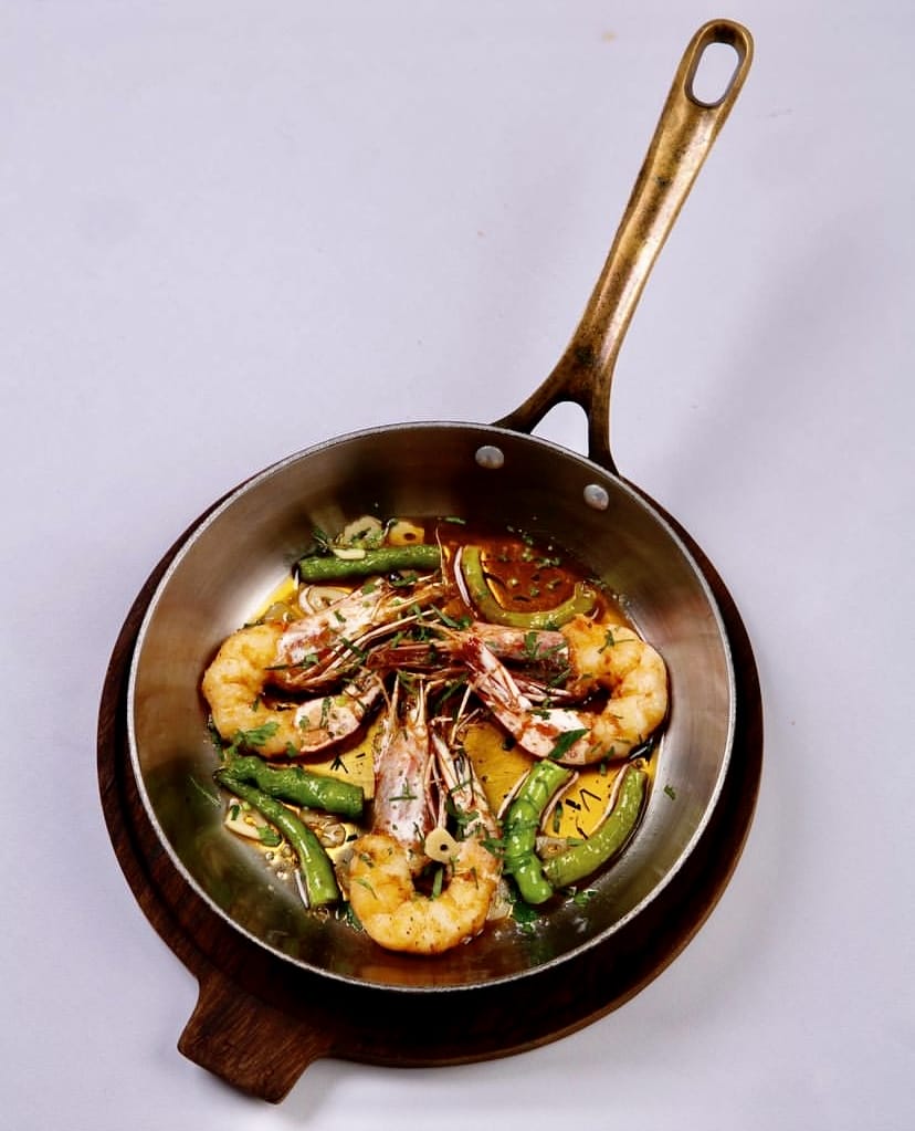 İstanbul'un en iyi teraslı mekânlarında keyifli bir gastronomik deneyimi çağrıştıran bu görselde, bronz saplı parlak metal bir tavada sarımsak ve yeşil biberlerle harmanlanmış, iştah açıcı karidesler ahşap bir sunum tahtası üzerinde özenle servis ediliyor.
