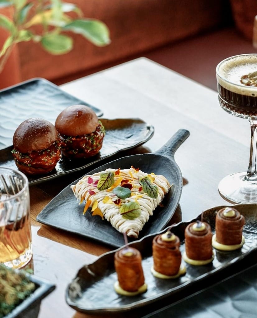 Şık bir masada, üzerinde yeşillik ve çiçeklerle süslenmiş üçgen şeklinde bir tatlı, iki mini burger, kremalı silindirik atıştırmalıklar ile köpüklü koyu renk bir kokteyl ve amber rengi bir içeceğin özenle sunulduğu bu görüntü, İstanbul'un en iyi teraslı mekanlarında sunulan yaratıcı lezzetleri ve imza kokteylleri vurguluyor.