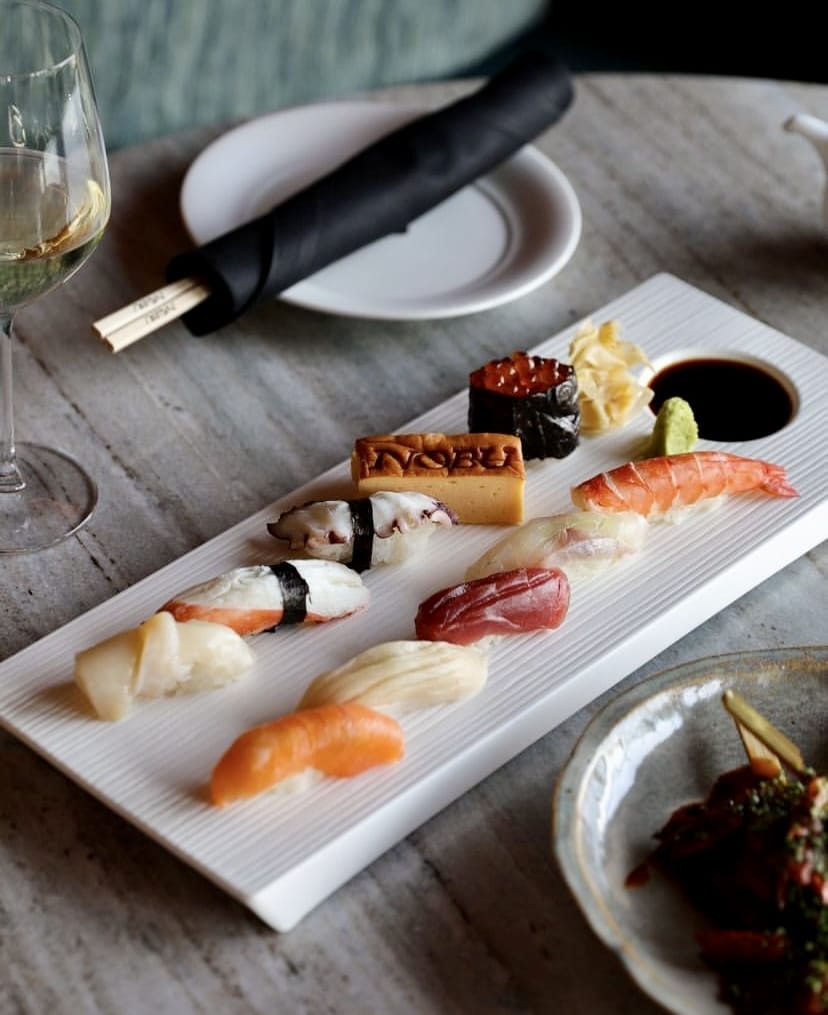 Beyaz, çizgili dikdörtgen bir tabakta sunulan; somon, orkinos, karides, ahtapot nigiri, tamago ve ikura gunkandan oluşan çeşitli sushi çeşitleri, yanında soya sosu, wasabi ve zencefil turşusu ile arka planda bir kadeh beyaz şarap ve yemek çubukları, İstanbul'un en popüler sushi restoranlarının özenli ve taze lezzetlerini yansıtıyor.