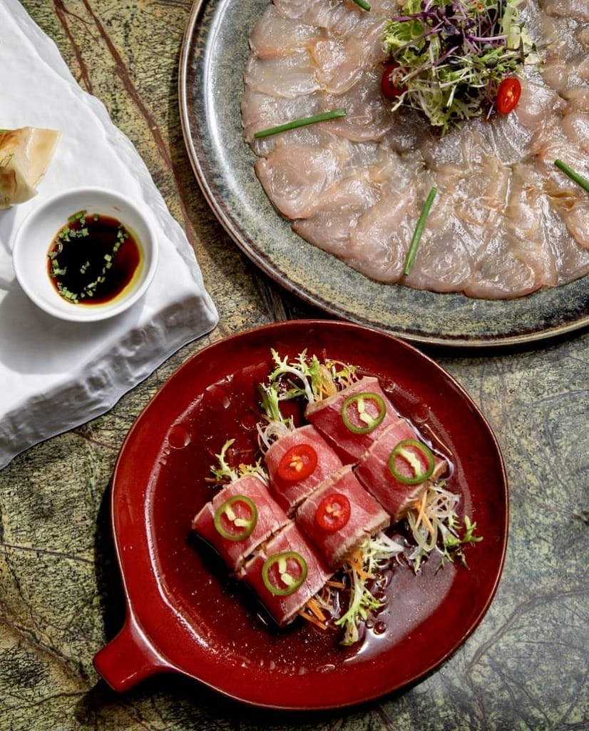 Mermer bir yüzeyde şıkça sunulmuş, ince dilimlenmiş balık sashimi'si, taze yeşillikler ve kırmızı biber dilimleriyle süslenmiş büyük bir tabak ile, yanında jalapeño ve kırmızı biber halkalarıyla zenginleştirilmiş, sebzeli özel tuna sushi roll'larının bulunduğu parlak kırmızı bir tabak, İstanbul'un en iyi sushi restoranlarında deneyimlenebilecek modern Uzak Doğu lezzetlerinin malzeme kalitesini ve şef dokunuşunu vurguluyor.