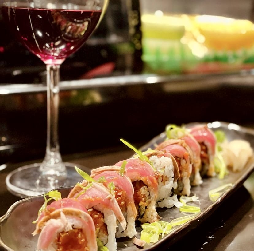 Koyu renkli bir tabakta özenle sunulan, üzeri hafifçe mühürlenmiş kırmızı et veya ton balığı ile kaplı, taze yeşil soğanlarla süslenmiş lezzetli sushi roll'ları, İstanbul'un popüler sushi restoranlarında tadına bakılabilecek modern Uzak Doğu lezzetlerini ve rafine sunumları kırmızı şarap eşliğinde sergiliyor.