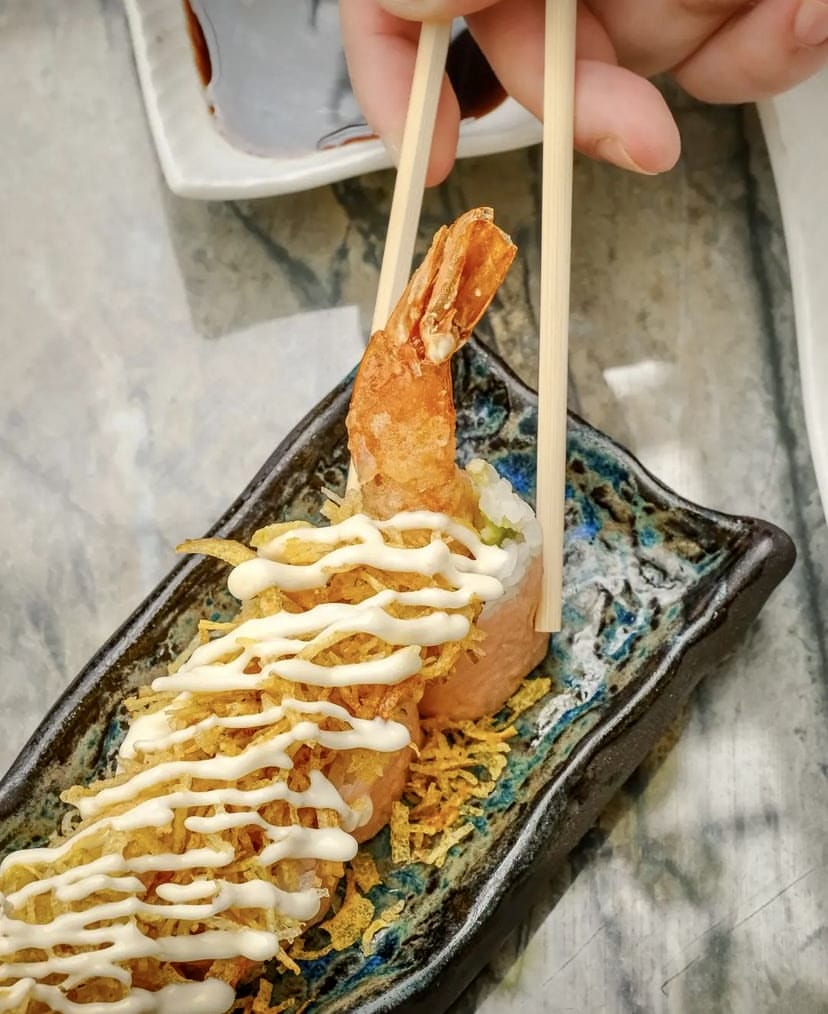 İstanbul'un önde gelen sushi restoranlarında sunulan, üzerinde çıtır tempura karides, mayonez sosu ve kızarmış çıtır garnitürler bulunan, somonla sarılmış özel bir sushi roll'un yemek çubuklarıyla alınırken çekilmiş yakın plan fotoğrafı, şehrin rafine Uzak Doğu mutfağındaki tazeliği ve imza lezzetlerini vurguluyor.