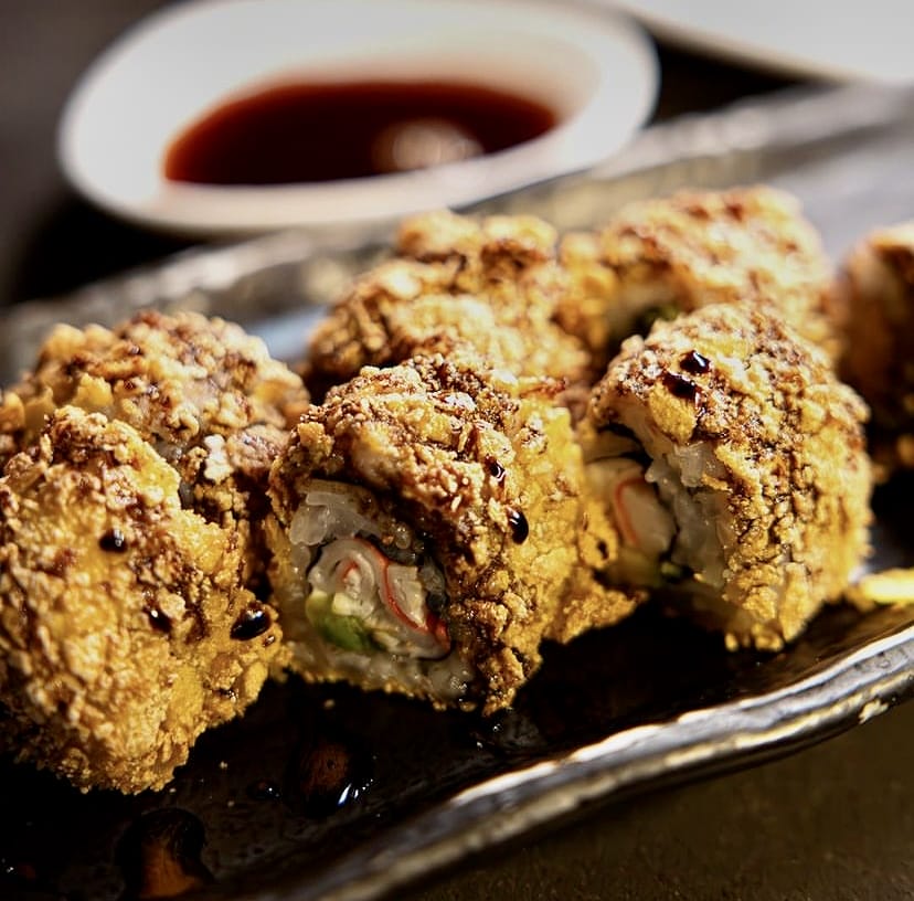 Altın renginde, çıtır dış kaplamalı, üzerine koyu renkli sos gezdirilmiş ve içi yengeç surimi ile avokado içeren, İstanbul'un çeşitli sushi restoranlarında sunulan modern bir yorum olan iştah açıcı tempura roll'lar, arka planda sos kasesiyle bir bütün oluşturuyor.