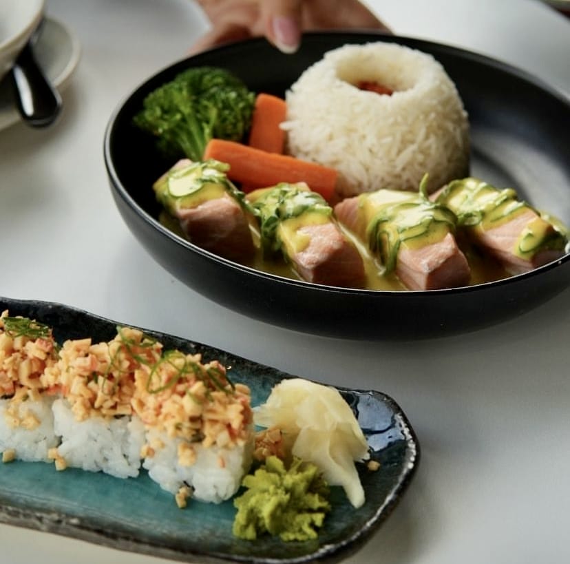Ön planda yengeç salatası benzeri karışımla zenginleştirilmiş dört adet iştah açıcı sushi rulo, taze wasabi ve zencefil turşusu ile birlikte zarif bir tabakta sunulurken, arka planda bir elin tuttuğu siyah kasede sarı soslu ve salatalık dilimli somon filetoları, pilav, brokoli ve havuç eşliğinde, İstanbul'un en popüler sushi restoranlarının sunduğu rafine Uzak Doğu lezzetlerini ve kaliteli sunumlarını vurguluyor.