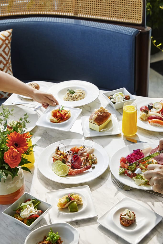 Bir masanın üzerinde, taze deniz ürünleri, suşi, salata ve tatlılarla dolu bol çeşitli bir brunch sofrası; bir yandan taze sıkılmış portakal suyu ve renkli çiçekler, diğer yandan yemek seçen misafirlerin elleriyle hareketli ve zarif bir ortam sunuyor.