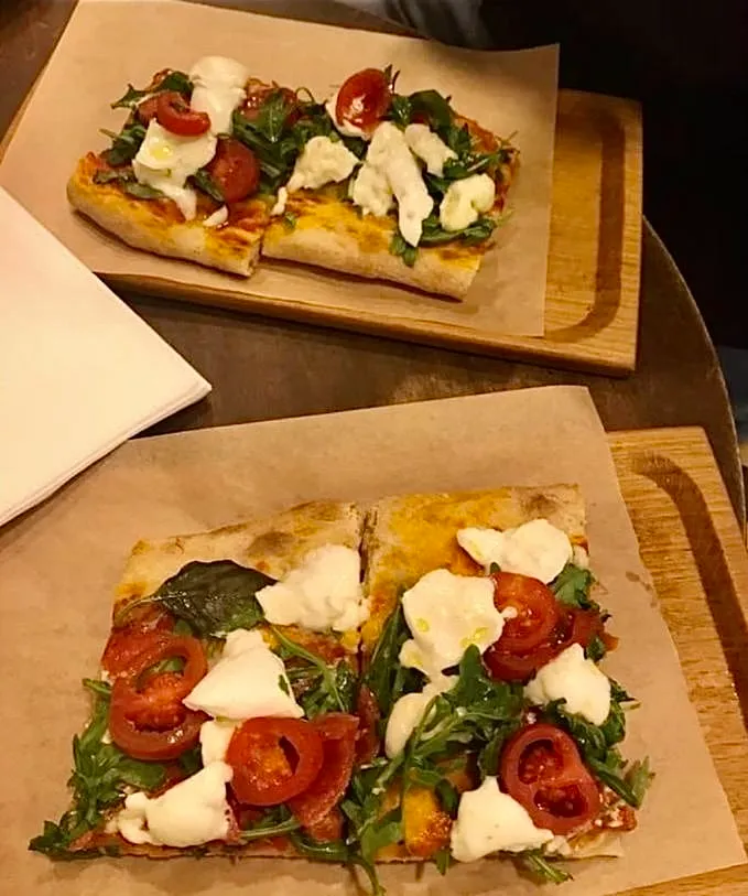 Masada, tahta servis tahtaları üzerinde kahverengi parşömen kağıtlarına yerleştirilmiş, roka yaprakları, dilimlenmiş kırmızı çeri domatesler ve bembeyaz taze mozzarella peyniri ile süslenmiş iki adet dikdörtgen dilim pizza, lezzetli bir İtalyan yemeği deneyimini çağrıştırıyor.
