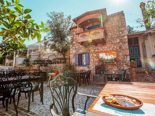 Güneşli havada misafirlerini ağırlayan, ahşap masada kremalı ve limon dilimli bir porsiyon lokma tatlısının öne çıktığı, taş duvarlı, ahşap balkonlu ve mavi pencereli geleneksel bir restoranın şirin açık hava avlusu.