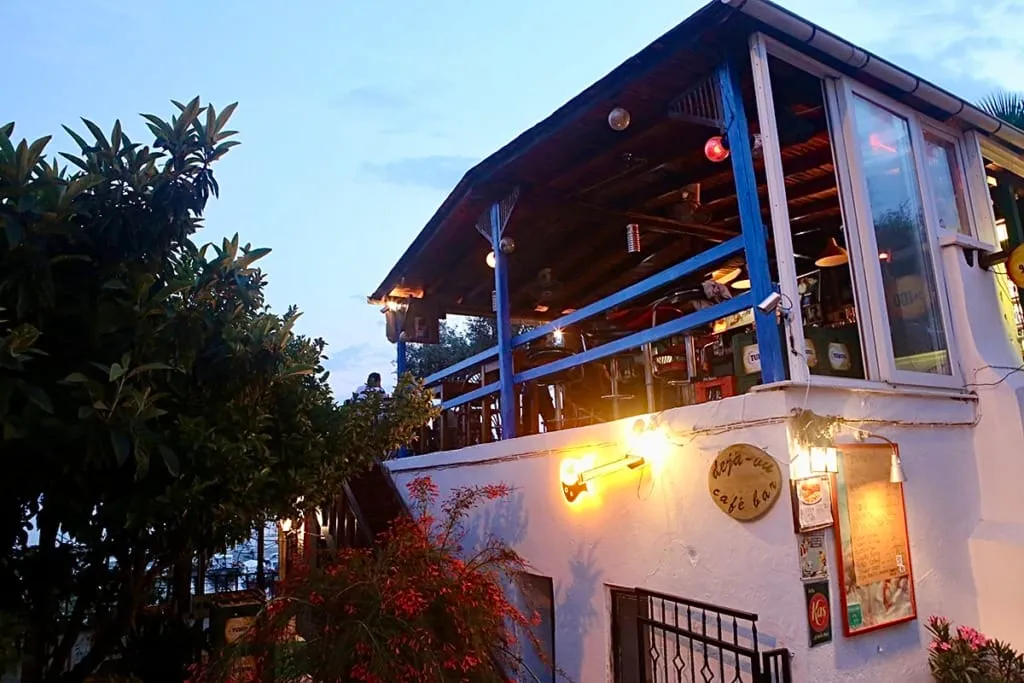 Akşamüzeri ışıklar altında, beyaz boyalı duvarlara ve ahşap çatılı üst kat terasına sahip, mavi korkuluklu canlı bir deja-vu cafe bar dışarıdan görülüyor; duvarındaki gitar şekilli ışıklandırma ve çevresindeki yeşil bitkilerle davetkar bir atmosfer sunuyor.
