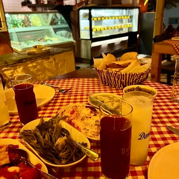 Kırmızı beyaz kareli bir masa örtüsü üzerinde, Demeti markalı buzlu rakı ve kırmızı bir içecek bardağının yanı sıra, deniz börülcesi, yoğurtlu ve domatesli mezelerin bulunduğu tabaklar ile bir ekmek sepeti yer alırken, arka planda bir restorana ait vitrinler ve hafifçe flu insanlar görülen, tipik bir Türk meyhanesi sofrası yansıması.