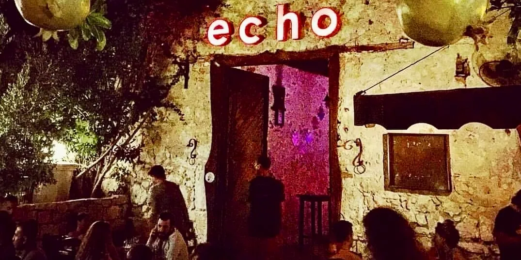 echo adlı mekanın kırmızı neon tabelasının altında, eski taş duvarlı bir binanın geniş açık kapısından içeri sızan mor ışığın aydınlattığı, ağaçlar ve loş aydınlatmalarla çevrili dış alanda sosyalleşen misafirler görülüyor.