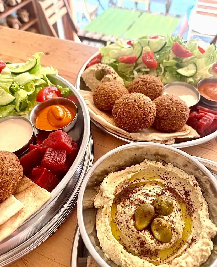 Ahşap bir masanın üzerinde, taze yeşil salata, kırmızı pancar küpleri ve çeşitli soslarla zenginleştirilmiş, üzerine zeytinyağı gezdirilmiş ve zeytinlerle süslenmiş büyük bir kase humusun da yer aldığı, çıtır falafeller ve pide ekmekleriyle dolu, gurme bir yemek tabağı görülüyor.
