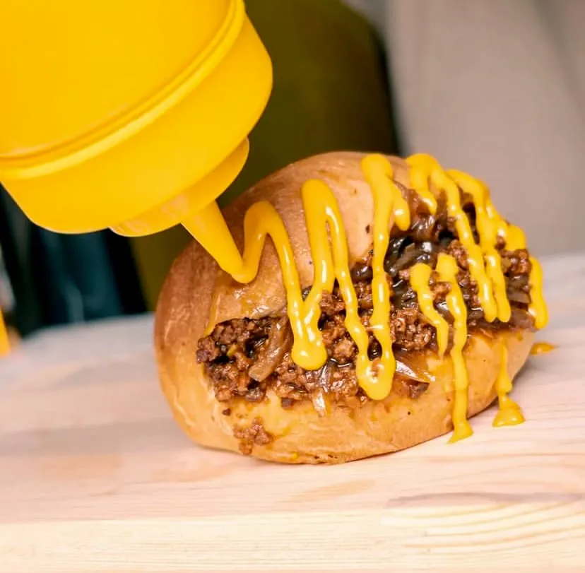 Açık renkli ahşap bir servis tahtası üzerinde duran, kıyma ve karamelize soğanlarla zengin bir şekilde doldurulmuş iştah açıcı bir hot dog sandviçin üzerine, sarı bir sos şişesinden akıcı bir şekilde parlak sarı bir sosun, muhtemelen hardal veya eritilmiş peynir sosunun, özenle döküldüğü yakın bir çekim.