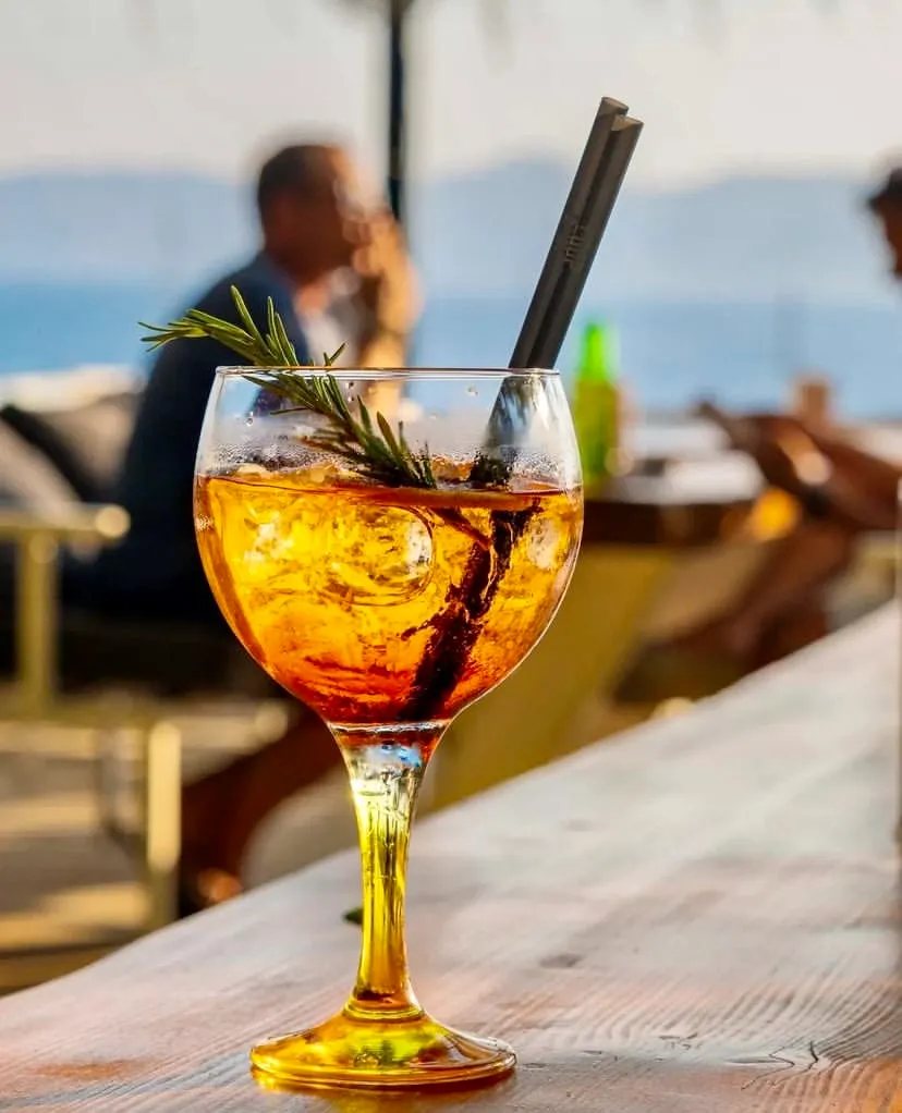 Kaş'taki bir tatil gününü çağrıştıran bu canlı karede, ahşap bir masanın üzerinde duran turuncu renkli kokteyl, içindeki buz küpleri, taze biberiye dalı ve iki şık koyu pipetle dikkat çekiyor; arka planda bulanık deniz ve rahatlayan figürler huzurlu bir atmosfer sunuyor.