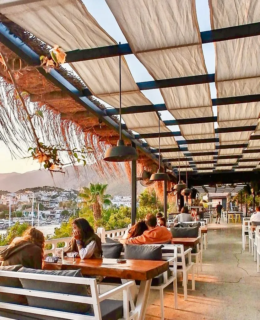 Kaş'ın büyüleyici manzarasına nazır bir açık hava restoranında, bej renkli kumaş tenteler ve sarkıt aydınlatmaların altında, ahşap masalarda oturan misafirler, gün batımının yumuşak ışıklarıyla aydınlanan denizi ve tepeleri izleyerek sakin bir akşamın keyfini çıkarıyor.