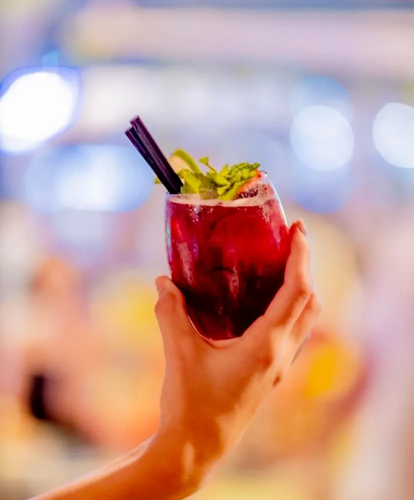 Kaş'ta keyifli bir akşamda Mavi Bar'ın hareketli atmosferini yansıtan bu karede, bir el nane, limon dilimi ve iki siyah pipetle süslenmiş, canlı kırmızı tonlarda ferahlatıcı bir kokteyl bardağını havaya kaldırırken arka plan bulanık ışıklarla dolu.