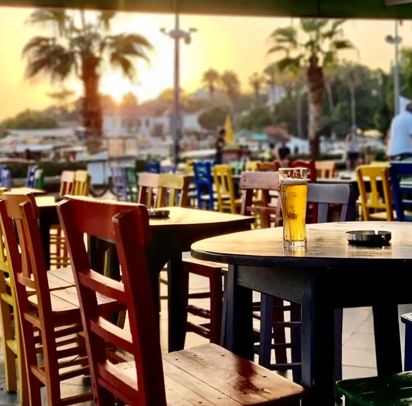 Kaş'taki Mavi Bar'ın dış mekanında gün batımında çekilen bu karede, masada köpüklü bira bardağı ve küllük görülürken, etrafta canlı renklerde sandalyeler ve palmiye ağaçları ile sıcak bir akşam atmosferi hâkimdir.