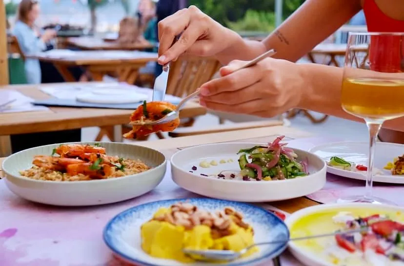 Açık hava bir restoranda, bir kişi çatal ve bıçak kullanarak karidesli, pilavlı bir mezeden kendi tabağına servis yaparken, masada salata ve püre benzeri rengarenk farklı mezeler ile bir kadeh içecek de görünüyor, Mezeteryan konseptini yansıtan zengin bir yemek deneyimi sunuyor.