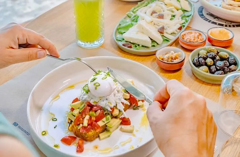 Bir kişinin elleri, ortasında haşlanmış yumurta, avokado ve domatesli, altında kızarmış ekmek veya fritür bulunan modern bir tabağı bıçak ve çatalla keserken görülüyor; arka planda yeşil bir içecek, çeşitli peynirler, zeytinler ve soslardan oluşan zengin bir kahvaltı sofrası yer alıyor.