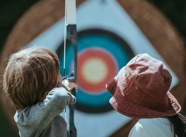 Bir çocuk, yanında şapkalı başka bir çocukla birlikte, yay ve ok kullanarak renkli bir okçuluk hedefine odaklanmış bir şekilde nişan alıyor; bu görsel, okçuluk sporunun hem konsantrasyon hem de kişisel gelişim için faydalarını vurguluyor.
