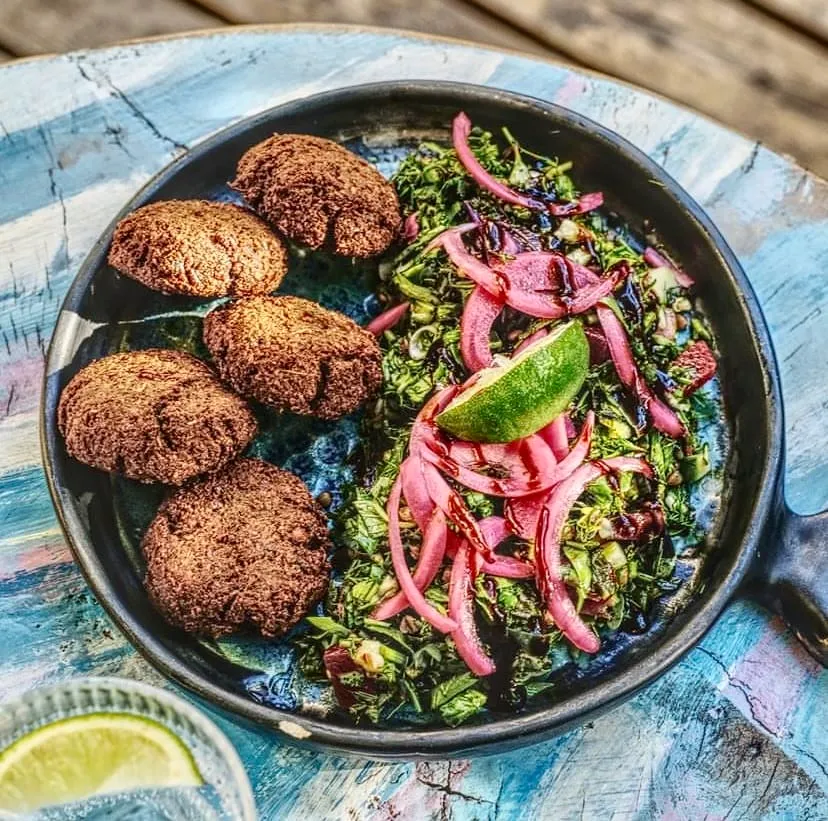 Kaş'taki Pells restoranında servis edilen bu tabakta, beş adet altın rengi çıtır falafel köftesi, taze yeşil otlar ve ince kıyılmış sebzelerden oluşan salata, üzerine parlak pembe soğan turşusu, koyu renkli bir sos ve bir dilim misket limonu eklenerek sunulan zengin ve lezzetli bir öğün görülmektedir.