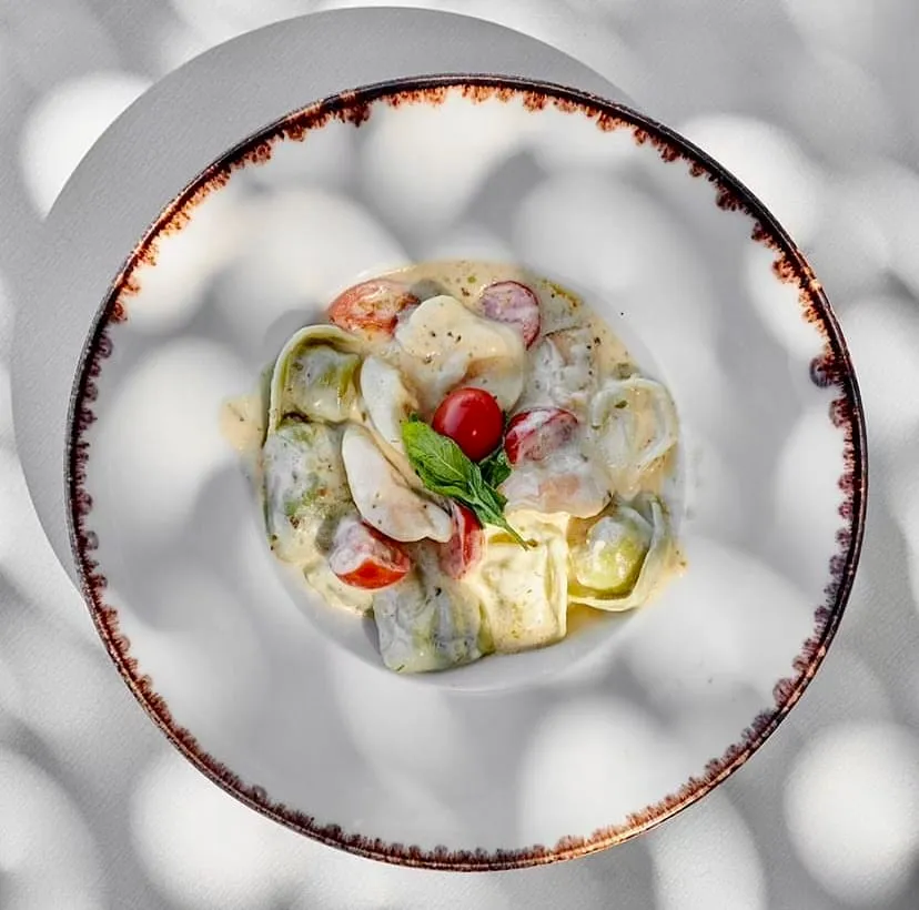 Şık, kahverengi kenarlı beyaz bir kasede servis edilen, yeşil ve beyaz tortellini veya ravioli parçacıklarından oluşan kremalı makarna yemeği, taze kiraz domatesleri ve üzerinde tek bir fesleğen yaprağı ile süslenmiş olup, yemek masası yüzeyine düşen dantelli gölgeler eşliğinde Sarnıç Plajı'nda keyifli bir öğünü yansıtmaktadır.
