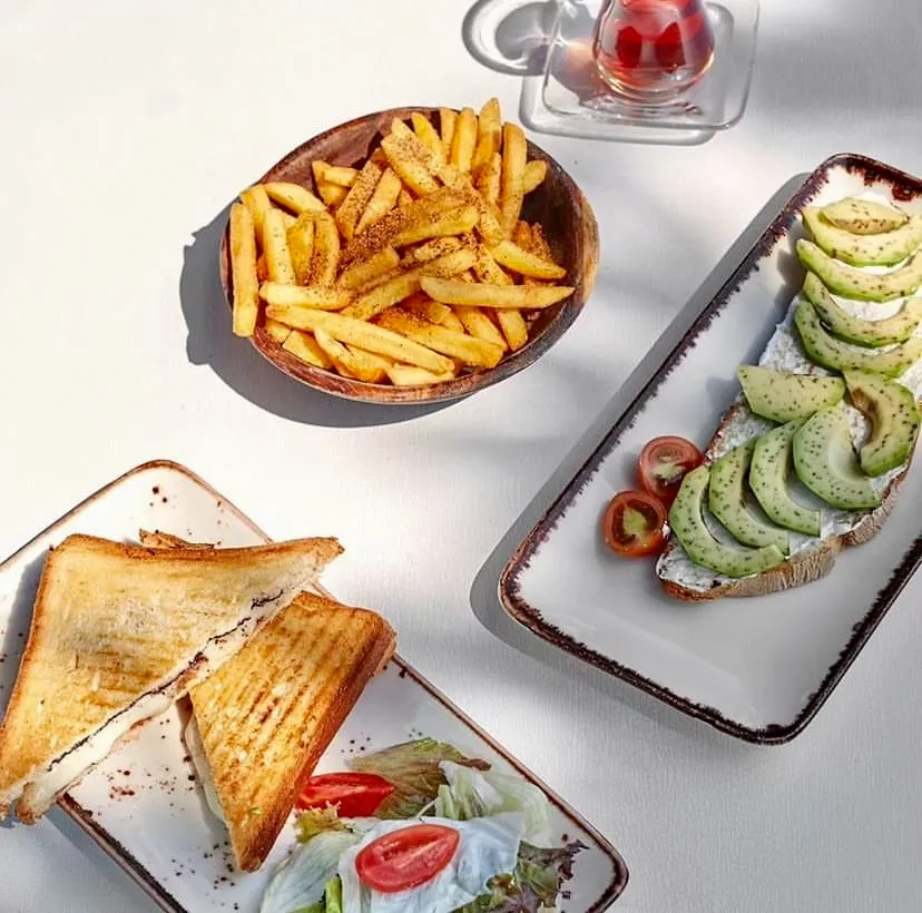 Sarnıç'ta güneşli bir masada, baharatlı patates kızartması dolu ahşap kase, krem peynirli ve avokado dilimli ekmek, yanında domatesli salatayla servis edilen tost ve bir bardak demli çay keyifli bir öğünü sergiliyor.