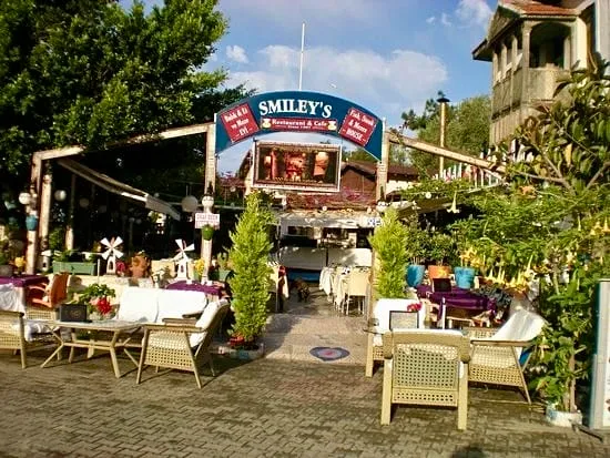 Smiley's Restaurant & Cafe'nin Arnavut kaldırımlı bir avluda yer alan, beyaz örtülü masalar ve hasır sandalyelerle dolu, yemyeşil ağaçlar ve sarı çiçekli bitkilerle çevrili, kısmen ahşap pergolalı açık hava yemek alanı gösterilmektedir; yukarıda ise balık şeklindeki tabelada restoranın balık, biftek ve daha fazlası sunduğu vurgulanmaktadır.