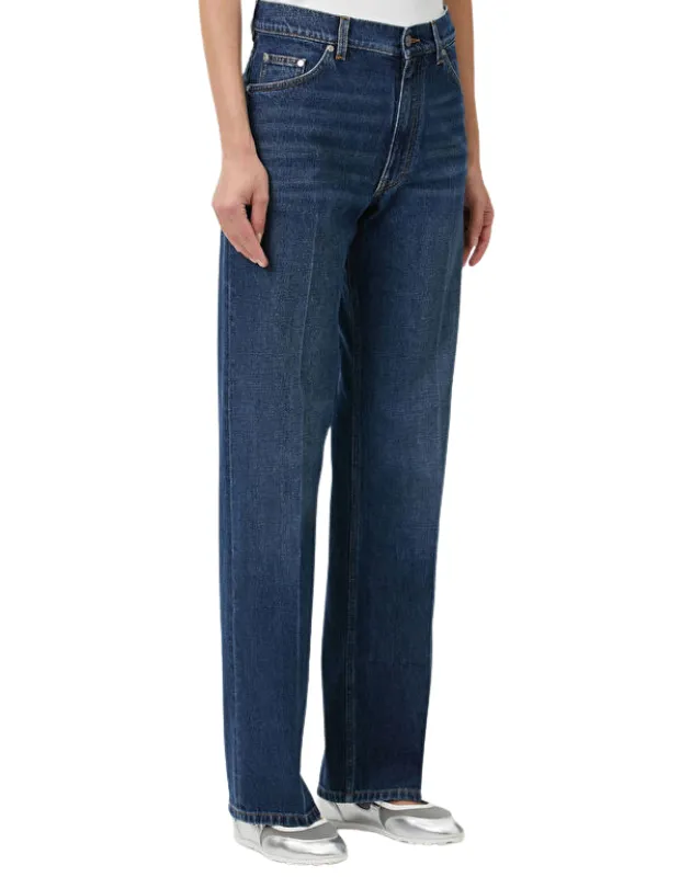 Kapsül gardırobun temelini oluşturan, her stile uyum sağlayan ve modası geçmeyen zamansız parçalardan biri olan koyu mavi straight leg jean pantolon, beyaz bir üst ve gümüş renkli düz ayakkabılarla kombinlenmiş olarak, beyaz stüdyo arka planı önünde ayakta duran bir kişinin bacaklarını gösteriyor.