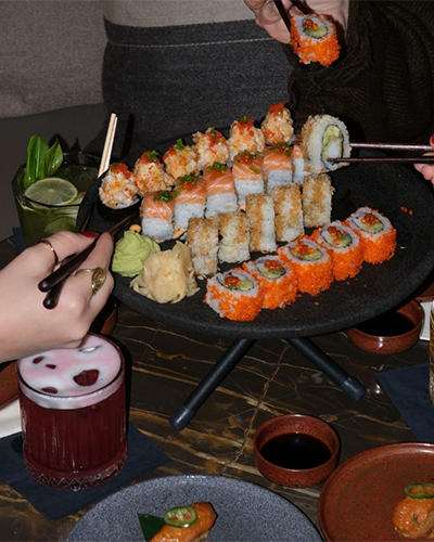 İstanbul'un önde gelen modern sushi duraklarından birinde, üzerinde turuncu tobiko kaplı, somonlu ve yengeçli gibi çeşitli sushi roll'larının sunulduğu siyah katlı servis tabağı etrafında, taze wasabi ve zencefil eşliğinde keyifli bir Uzak Doğu lezzet deneyimi paylaşılıyor.