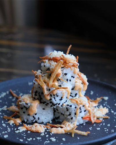 Koyu renkli bir tabakta pirinç ve siyah susam taneleriyle kaplı, üzeri özel turuncu sos gezdirilmiş ve çıtır kızarmış parçacıklarla süslenmiş, İstanbul'un en iyi sushi restoranlarından birinin modern dokunuşlarla hazırladığı yaratıcı sushi roll'ları dikkat çekiyor.