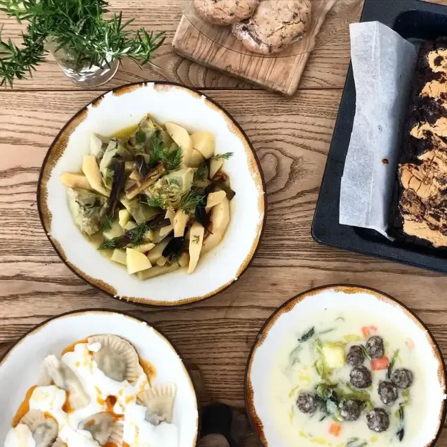 Bir ahşap masa üzerinde, enginarlı patates yemeği, yoğurtlu mantı, sebzeli köfteli çorba gibi geleneksel lezzetlerin yanı sıra kurabiye ve brownie gibi tatlıların da bulunduğu zengin bir öğün sergileniyor.